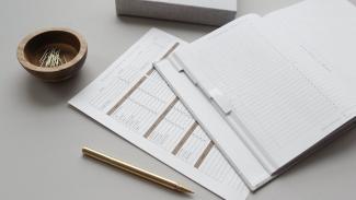 A desk with office supplies on it. In the middle of the desk is an open journal with a heading that reads "Finance Tracker". Under the journal is a piece of paper that reads "Monthly Budget Overview", and has several columns on it for tracking expenses.